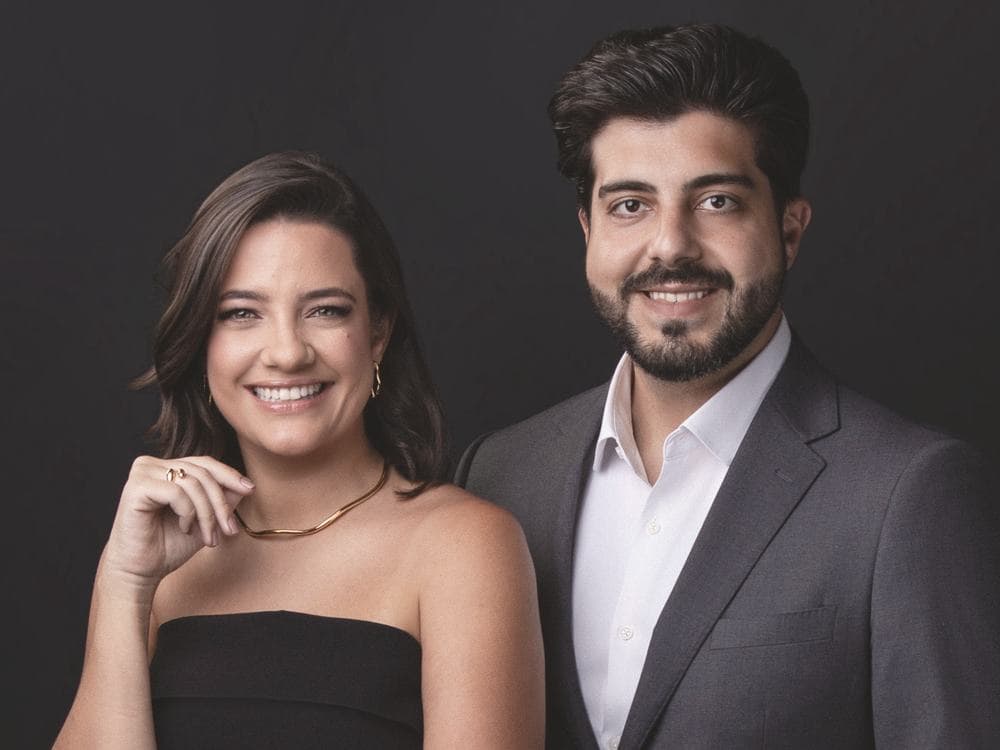 Dra. Nayara Curti e Dr. Renan Gerbelli, lado a lado e sorrindo, em retrato de estúdio com fundo escuro